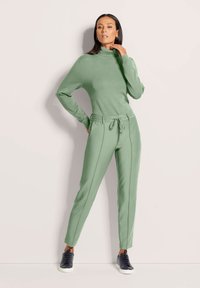 Vrouw staat met één hand op de heup en de andere hand tegen haar nek, gekleed in een groene coltrui en bijpassende jogpants met touwtje.