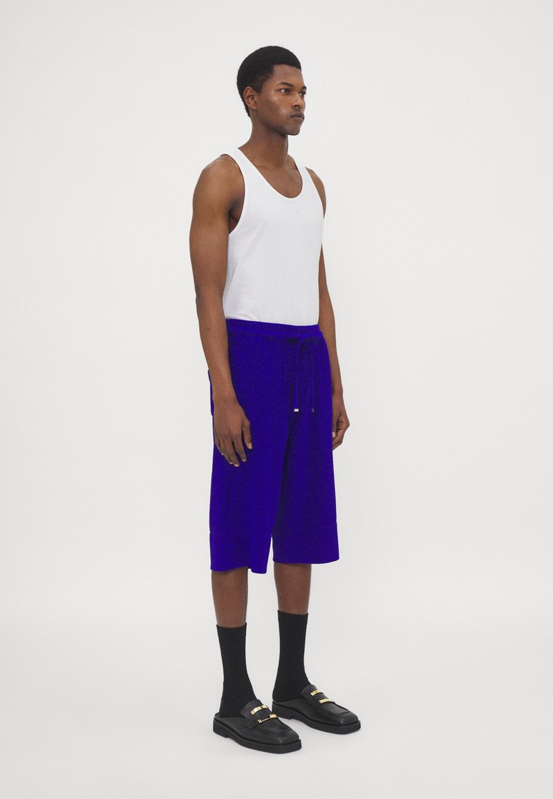 Paarse gebreide shorts met een taillekoord, gecombineerd met een wit tanktop, zwarte sokken en zwarte instappers met gouden accenten.