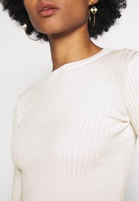 Femme aux cheveux bouclés foncés porte de grandes boucles d’oreilles créoles dorées avec des perles et un top blanc en maille côtelée à manches longues.