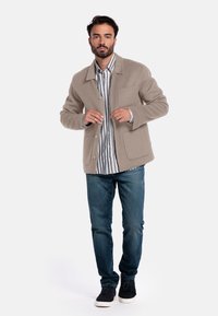 Veste en mélange de laine beige avec un col, fermeture à boutons sur le devant et deux poches, portée sur une chemise rayée blanche et bleue et un jean bleu.