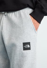 Person iført grå sweatpants med The North Face-logo på låret og en sort langærmet trøje, hånd i lommen.