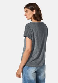 Camiseta gris de manga corta con un corte relajado, mangas enrolladas y acentos de botón en el hombro, situada sobre un fondo claro.