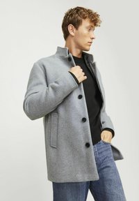 Abrigo de lana gris con cuello alzado, cierre de botones, bolsillos laterales y un corte holgado, combinado con un suéter negro y unos jeans azules.
