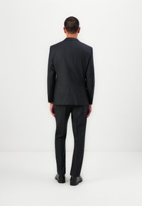 Dunkelgrauer Anzug mit tailliertem Jacket und schmal zulaufenden Hosen, aus glattem Stoff gefertigt. Schlichte Ausführung, minimale Details, getragen mit schwarzen Schuhen.
