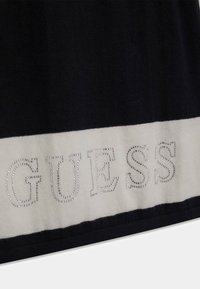 Mörkblått tyg med en vit rand som har ordet "GUESS" i silverfärgade rhinestones. Slät textur med kontrasterande färger.