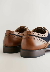 Pruunid nahast kingad, millel on sametise pealsega, brogue detailid ja ümar nina. Nahkne konts ja kummist tald tagavad vastupidavuse.