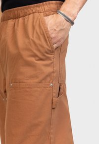 Brown cargo pants with an elastic waistband, side pockets, and snap buttons. Fabric has a smooth texture with a matte finish.