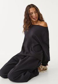 Černá oversized mikina s designem bez ramene, měkkou texturou a žebrovanými manžetami, v kombinaci s tmavými volnými kalhotami a světlými teniskami.