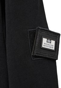 Tissu noir avec un patch rectangulaire noir portant l'inscription "WEEKEND OFFENDER", cousu avec du Velcro sur la surface.