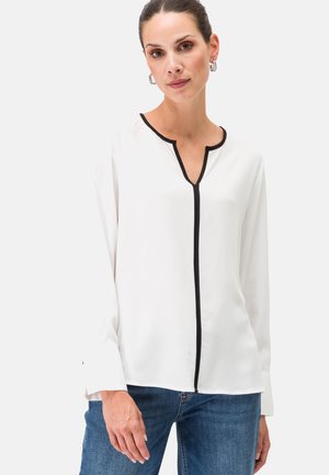 Blouse blanche avec un décolleté en V et des liserés noirs, manches longues avec des poignets à boutons, en tissu lisse. Portée avec un jean bleu.