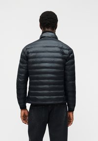 Άνδρας με γυαλιστερό μαύρο καπιτονέ μπουφάν και μαύρο τζιν, στέκεται γυρισμένος πλάτη σε απλό λευκό φόντο.