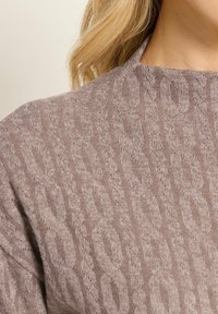 Primo piano di spalla e collo che indossa un maglione a maglia color mauve con un motivo leopardato testurizzato e lunghi capelli biondi.