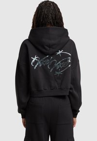 Felpa nera crop con un design grafico argentato che presenta stelle e testo scritto a mano sul retro. Materiale morbido e vestibilità rilassata.