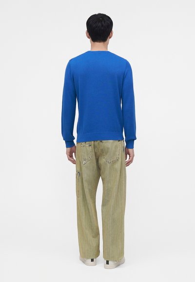 Homme de dos portant une chemise bleue à manches longues, un pantalon beige ample avec des poches arrière et des baskets blanches sur un fond uni.