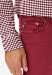 Jeans di denim rossi con un taglio classico, abbinati a una camicia a quadri bordeaux e bianca con polsini con bottoni. Piccolo logo sulla tasca porta orologio.