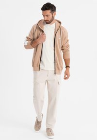 Beige zip-up huvtröja med mjuk textur, i kombination med krämfärgade cargobyxor och ljusa sneakers, med detaljerad sömnad och fickor.