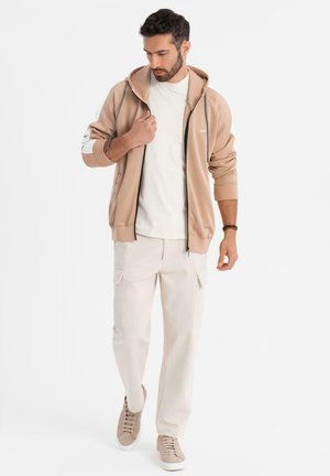Beige Kapuzenpullover mit Reißverschluss aus weichem Material, kombiniert mit cremefarbenen Cargo-Hosen und hellen Sneakers, die über detaillierte Nähte und Taschen verfügen.
