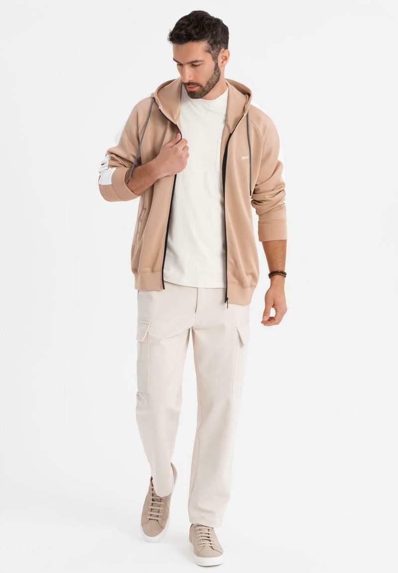 Beige zip-up huvtröja med mjuk textur, i kombination med krämfärgade cargobyxor och ljusa sneakers, med detaljerad sömnad och fickor.
