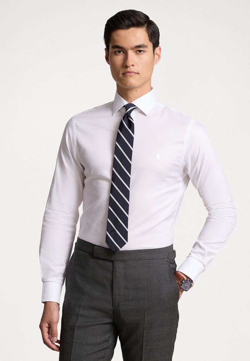 Chemise blanche à la coupe ajustée, dotée d'un petit logo sur la poitrine et accompagnée d'une cravate rayée bleue marine ; assortie à un pantalon gris sur mesure.