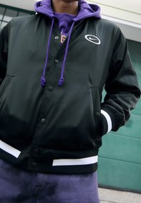 Černá nylonová bomber bunda s bílými pruhy na lemu a manžetách. Obsahuje velké logo a vrstvenou fialovou mikinu pod tím.