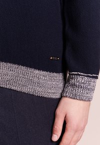 Pull tricoté bleu marine avec poignets et ourlet côtelés gris et blancs, montrant la main d'une personne reposant à côté de la manche.