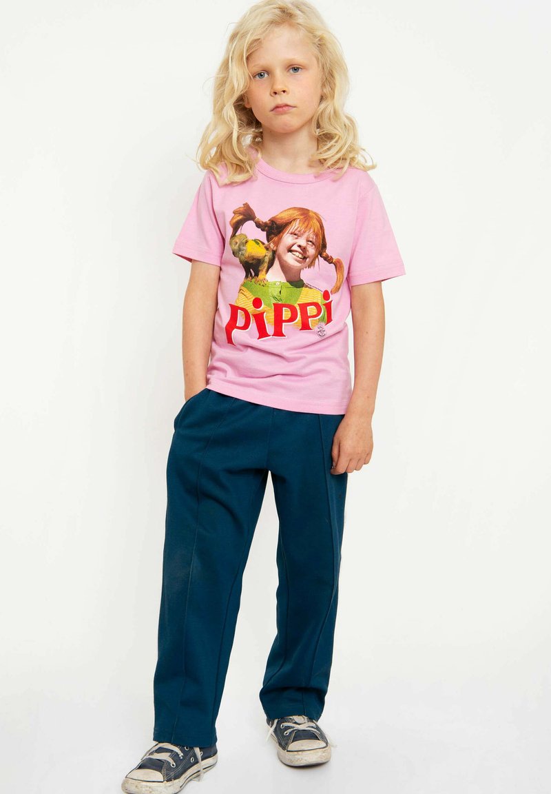 Rosa T-Shirt mit "PIPPI" Aufdruck und einer bunten Grafik, kombiniert mit dunkelblauen Hosen und abgewetzten Sneakers. Lange, gewellte blonde Haare.