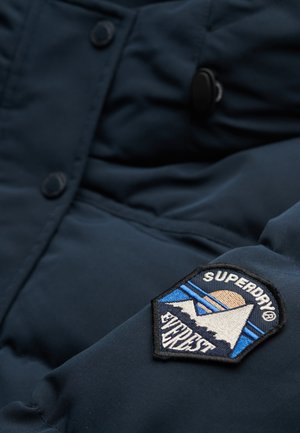 Marineblauwe gewatteerde jas met een zwarte knop, een gladde textuur en een geborduurd Superdry-logo met een berg- en zonontwerp.