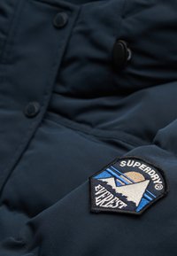 Granatowa puchowa kurtka z fakturowaną tkaniną, zaokrąglonym kołnierzem oraz naszywką z haftowanym wzorem góry i słońca po lewej stronie.