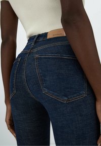 Jeans de mezclilla azul oscuro con un diseño ajustado, que presentan dos bolsillos traseros, costuras en contraste y una etiqueta de cuero marrón en la cinturilla.