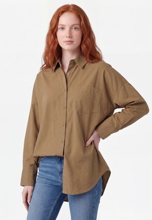Rødhåret kvinde iført en løs brun skjorte med knapper og blå jeans, stående med den ene hånd på hoften mod en ensfarvet baggrund.