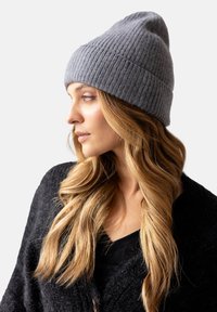 Szary, prążkowany beanie o stożkowym kształcie, wykonany z miękkiego materiału dzianinowego. Noszony z ciemnym kardiganem, z długimi falującymi włosami opadającymi na jedną stronę.