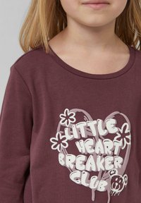 Bordowy longsleeve z okrągłym dekoltem, na którym znajduje się biały nadruk przedstawiający serce, kwiaty i misia. Miękka tekstura, casualowy styl.