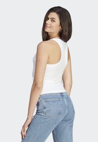 Femme aux cheveux noirs raides portant un débardeur blanc sans manches et un jean bleu taille haute, se posant légèrement tournée vers l'arrière.