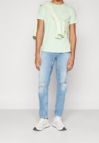 Světle zelené bavlněné tričko, na kterém je přes ramena přehozený odpovídající svetr. Oblečené jsou vybledlé modré džíny s roztržením. Bílá teniska dotváří vzhled.