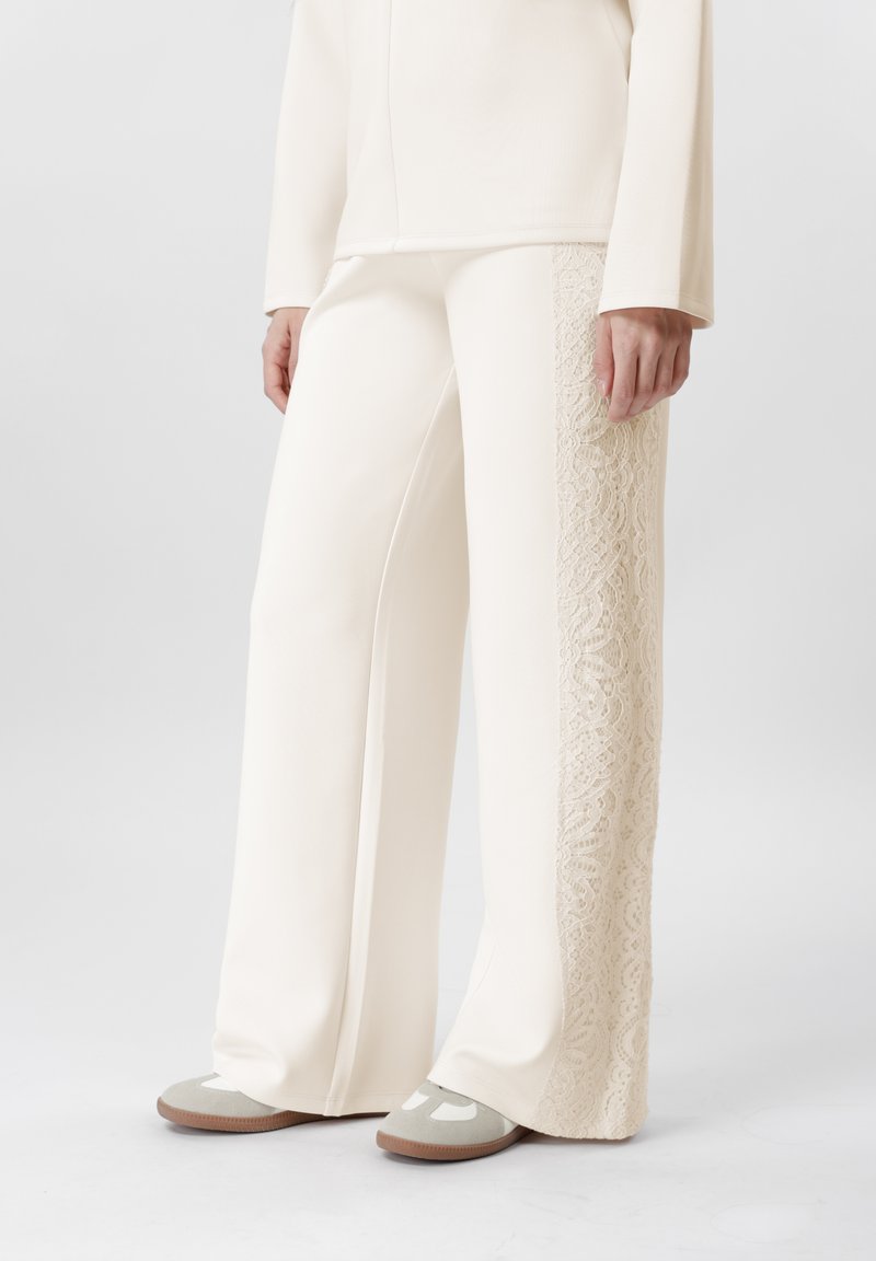 Persona vistiendo pantalones anchos color crema con panel de encaje en el lado exterior, camiseta de manga larga color crema y zapatos planos beige, de pie sobre un suelo gris.