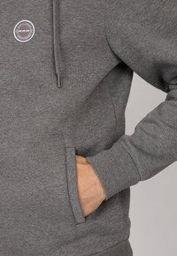 Sweat à capuche gris avec une texture douce, poche kangourou et un patch logo rond sur la poitrine. Comprend une capuche avec cordon de serrage et des poignets côtelés.