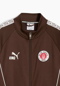 Chaqueta marrón con cremallera y cuello alto, acentos en blanco y logo en el pecho. Presenta rayas texturizadas a lo largo de los hombros. Tejido ligero.