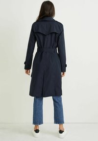 Granatowy trencz z zapięciem na guziki z tyłu i paskiem, z długimi rękawami i długością do kolan, zestawiony z niebieskimi dżinsami i białymi butami.