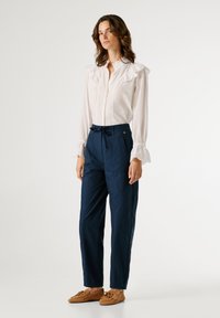 Λευκό πουκάμισο μακρυμάνικο με γιακά με βολάν, συνδυασμένο με παντελόνια σε σκούρο μπλε χρώμα με ψηλή μέση. Καφέ μοκασίνια ολοκληρώνουν το ντύσιμο. Απλός σχεδιασμός.