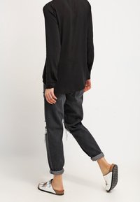 Person wearing black long-sleeve shirt, rolled-up dark jeans, and white slip-on sandals walking against a plain light background.