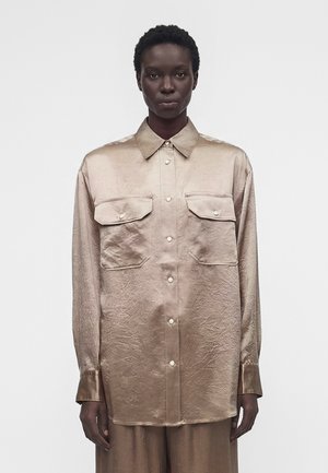 Osoba ubrana w beżową, satynową koszulę zapinaną na guziki z dwoma kieszeniami na piersi, stojąca na jednolitym jasnoszarym tle.