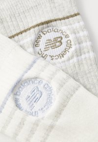 Calcetines New Balance en crema claro con tejido texturizado, que presentan patrones de rayas claras y oscuras y logotipos bordados en dorado y azul claro.