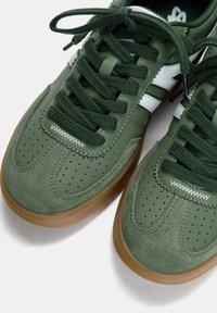Baskets vertes présentant un mélange de cuir et de daim, un design perforé, une semelle en caoutchouc et un système de laçage avec des lacets vert foncé.
