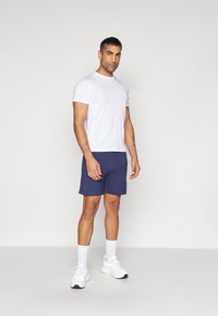 Camiseta blanca de cuello redondo con un logotipo sutil, combinada con pantalones cortos navy que tienen un pequeño logotipo bordado, y zapatillas deportivas blancas con detalles visibles.