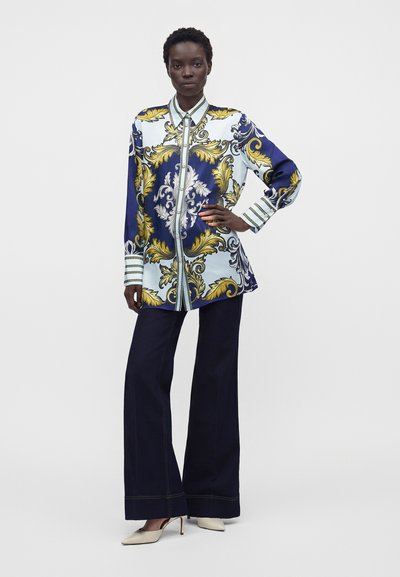 Femme debout, la main sur la hanche, portant une longue chemise à motifs avec des dessins dorés et bleus, un pantalon large et foncé, et des talons blancs.