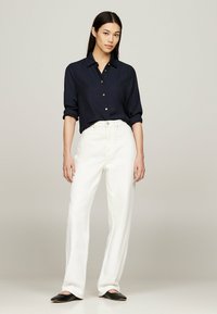 Camicia blu scuro con bottoni, maniche arrotolate, abbinata a pantaloni bianchi a gamba larga e scarpe nere slip-on, caratterizzata da una texture di tessuto liscia.