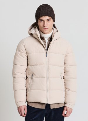 Beige Stepp-Steppjacke mit Kapuze und Reißverschlusstaschen, getragen über einem Hemd mit Kragen und dunkler Hose. Braune, gerippte Mütze.