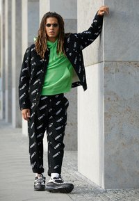 Černý outfit skládající se z bundy a kalhot s bílým logem "OBEY", doplněný jasně zelenou mikinou a černobílými teniskami.