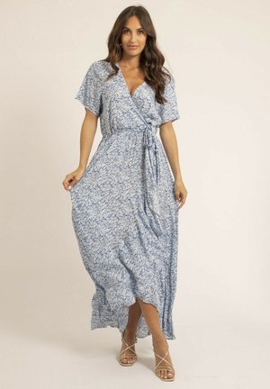 Femme portant une robe maxi portefeuille à fleurs bleues claires, tenant la jupe d'une main, debout dans un décor neutre avec des talons dorés à fines lanières.