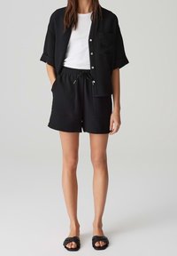 Černá strukturovaná košile s krátkým rukávem a kapsou, spárovaná s odpovídajícími černými kraťasy s elastickým pasem a šňůrkou. Černé sandály.